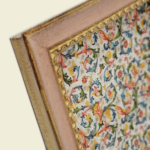 Può includere: Un'opera d'arte rettangolare incorniciata caratterizzata da un motivo denso e colorato di fiori e viticci vorticosi nei toni del rosso, blu, giallo e verde su sfondo bianco, racchiusa in una cornice rosa chiaro con bordo interno dorato.
