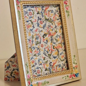 May include: A cream-coloured picture frame with a gold beaded inner border and floral accents. The frame holds patterned paper with a colourful, swirling design. The frame is propped up by a matching easel back.