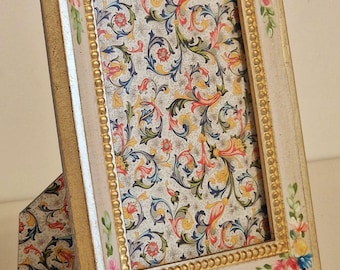 May include: A rectangular, cream-colored picture frame with a gold beaded inner border and floral accents. The frame holds a patterned paper with a colorful, swirling design. The frame is propped up by a matching easel back.