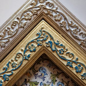 Puede incluir: Primer plano de dos marcos de fotos ornamentados. El marco superior es plateado con detalles florales. El marco inferior es dorado con detalles florales azules. Los marcos están en ángulo, revelando un fondo estampado con diseños florales azules y verdes.