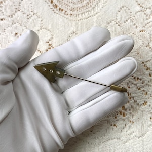 May include: A vintage arrow-shaped lapel pin. The pin features a brass-colored arrow with three small, round, clear embellishments. The pin has a long, thin, silver-colored shaft and a brass-colored end.