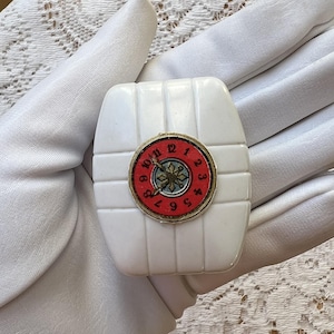 May include: A white, rectangular vintage compact with a red and gold clock face. The compact has a ribbed design and a small, circular clock with black numbers and a gold center. The clock face is surrounded by a gold rim.