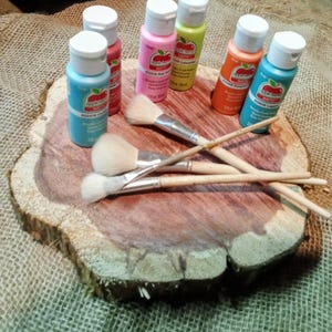 May include: A selection of acrylic paints in shades of blue, pink, and orange, in bottles with white lids. Paintbrushes with wooden handles and soft bristles are arranged on a wooden slice, indicating an art or craft project.