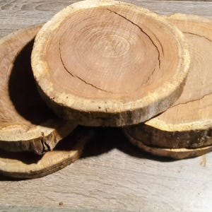 May include: A stack of four unfinished, round wooden slices with visible growth rings. The wood has a light brown color with a lighter-colored outer edge. The slices vary slightly in size and are stacked on a wooden surface.
