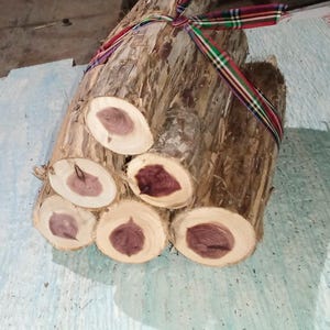 Peut inclure: Un fagot de bûches de cèdre, coupées pour révéler le cœur de bois rouge-violet. Les bûches sont liées avec un ruban à carreaux coloré. Le bois a un aspect naturel et rustique.