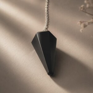 Könnte beinhalten: Ein facettierter, kegelförmiger schwarzer Anhänger, der an einer dünnen silberfarbenen Metallkette hängt. Der Anhänger hat eine glatte, matte Oberfläche mit scharfen geometrischen Kanten, die nach unten spitz zuläuft und eine dunkle Silhouette bildet.