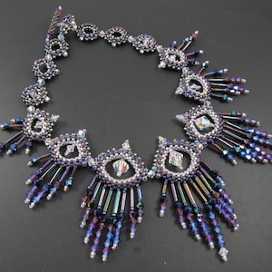 May include: A beaded necklace with a silver and iridescent purple color scheme. The necklace features a repeating pattern of teardrop shapes with a central crystal bead. The necklace has a fringe of beads hanging from the bottom.