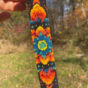 May include: A colorful beaded bracelet with a geometric pattern in shades of orange, yellow, blue, and green. The bracelet is made with small, round beads and has a black background. The bracelet is secured with metal clasps.