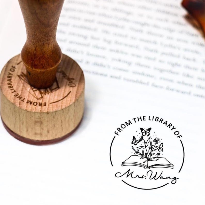 May include: A wooden library stamp with a brown handle and a circular stamp face. The stamp face reads "FROM THE LIBRARY OF" around a graphic of an open book with butterflies and flowers. The name "Mrs. Wang" is below the graphic.