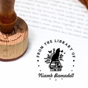 Puede incluir: Sello en blanco y negro con el texto "From the Library of Niamh Bamadell". El sello presenta una silueta de un gato sentado en una pila de libros con mariposas y hojas alrededor.