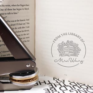 Puede incluir: Un sello de libro plateado con una banda dorada. El sello está impreso con el texto "From the Library of Mrs. Wang" y un diseño de un libro abierto con flores alrededor.