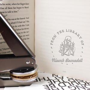 May include: A silver self-inking stamp with a gold band. The stamp features a skeleton reading a book with the text "From the Library of" and "Niamh Bamadell" in a circle around the skeleton.