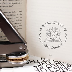 Puede incluir: Un sello de metal gris con un anillo dorado. El sello está diseñado para crear un ex libris personalizado con el texto "From the Library of Abby Sumner" y una ilustración en blanco y negro de un gato sentado en un libro abierto.