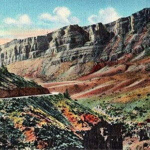 May include: Vintage postcard depicting a scenic mountain landscape with a winding road. The image showcases a canyon with layered rock formations in shades of brown, red, and green. A blue sky with clouds is visible above.