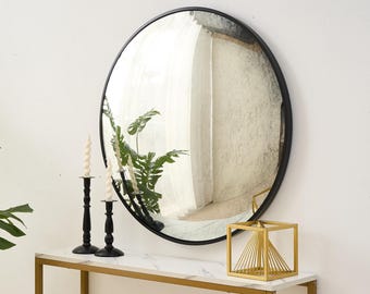 Pode incluir: Um espelho redondo grande com moldura preta está pendurado em uma parede branca. Abaixo, uma mesa com tampo de mármore branco e pernas douradas exibe castiçais pretos, velas em espiral e uma escultura geométrica dourada. Uma planta verde é visível.