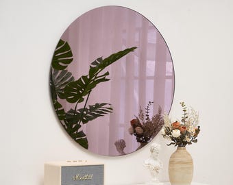 May include: A round, pink-tinted mirror hangs on a white wall, reflecting a room with plants and decor. Below, a cream-colored speaker with gold trim and a vase with dried flowers sit on a white table.