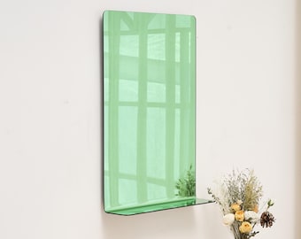 May include: A rectangular, green-tinted mirror with a black frame and a small shelf. The mirror is mounted on a white wall. The reflection shows a window with a grid pattern. A vase of flowers sits below the mirror.