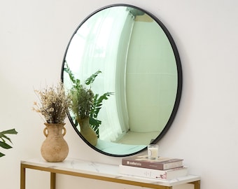 May include: A round black-framed mirror hangs above a marble-topped console table. The mirror reflects a bright room with a window. A vase of dried flowers and a vase of greenery sit on the table, along with books and a candle.