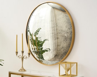 Pode incluir: Um espelho redondo com moldura dourada está pendurado em uma parede branca, refletindo uma sala. Um castiçal dourado com três velas e uma escultura geométrica dourada estão em uma mesa com tampo de mármore branco e pernas douradas.