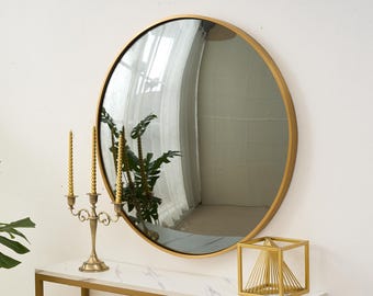 May include: A round gold-framed mirror hangs on a white wall. A gold candelabra with three candles, a gold geometric sculpture, and a plant sit on a white marble-topped table with a gold frame. The mirror reflects a window.