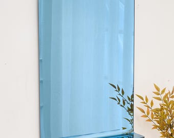 May include: A rectangular, blue-tinted mirror with a black frame and a small shelf at the bottom. The mirror is mounted on a white wall, with a few green and yellow plants in the background.