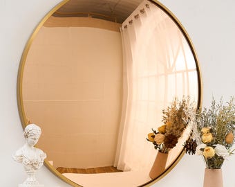 May include: A large round mirror with a gold frame and a rose gold tinted glass. The mirror is reflecting a white curtain and a bouquet of dried flowers in a vase.