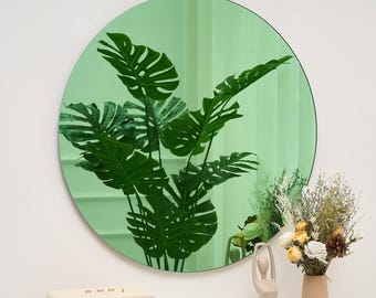 May include: A round, green-tinted mirror reflects a large green plant with split leaves. A small speaker, a sculpture of a person, and a vase of flowers are in front of the mirror. The mirror is mounted on a white wall.