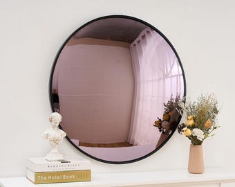 May include: A round, pink-tinted mirror with a black frame. The mirror is reflecting a room with white walls and a white curtain. The mirror is sitting on a white shelf with two books, a small white statue, and a vase with dried flowers.