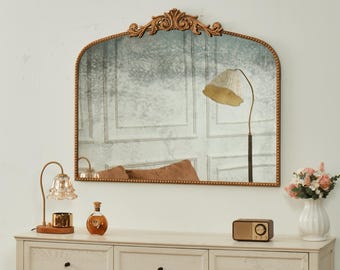 May include: An arched, antique-style mirror with a gold-colored frame and ornate detailing at the top. The mirror reflects a room with a floor lamp, a dresser, and decorative items, including a lamp, a bottle, a radio, and a vase of flowers.