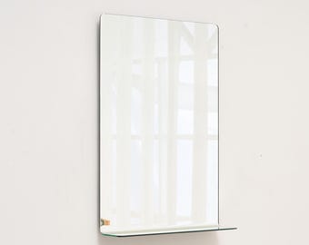May include: A rectangular wall mirror with a small shelf at the bottom. The mirror has rounded corners and a thin black frame. The reflection shows a window with white curtains. The mirror is mounted on a white wall.