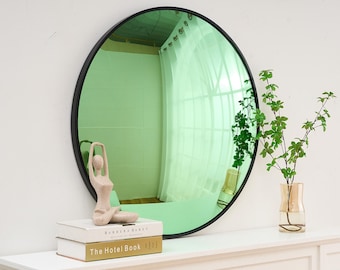 May include: A round green mirror with a black frame, hanging on a white wall. A small decorative statue of a person in a yoga pose is in front of the mirror, on top of two books. The books are titled "Barbara Barry" and "The Hotel Book".