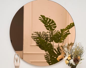 May include: A round, rose-gold mirror with a black frame. The mirror reflects a green leafy plant and a white wall. A small statue and a vase of flowers are in the foreground, creating a decorative and modern aesthetic.