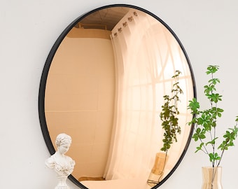 May include: A round, copper-toned mirror with a black frame. The mirror is reflecting a room with white curtains and a window. The mirror is sitting on a table with a book and a small statue.