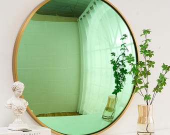 May include: A round, gold-framed mirror with a green, reflective surface. The mirror is hanging on a white wall and reflects a plant in a vase.
