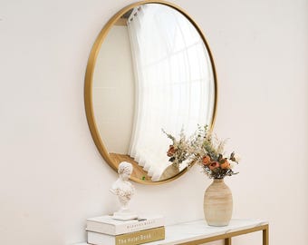 May include: A gold round mirror with a simple frame hangs on a white wall above a white marble console table. The table has a gold metal frame and is decorated with a white bust, two books, and a vase of dried flowers.