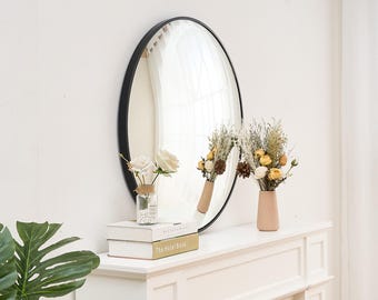 May include: A round black framed mirror with a white wall behind it. Two vases with dried flowers are on a white mantel with two books stacked on top of each other. The top book is titled "The Hotel Book".