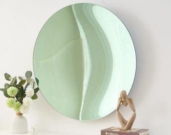 May include: A large, round, light green mirror with a unique, wavy design. The mirror is mounted on a white wall. A small sculpture of a person sitting with their head in their hands is on a stack of books.