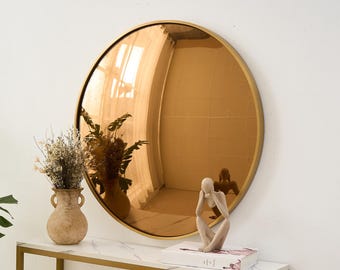 May include: A round, gold-framed mirror with a copper-toned reflective surface hangs on a white wall. The mirror reflects a room with natural light. A marble-topped console table with gold legs holds a vase of dried flowers, a vase of greenery, and a sculpture.