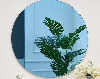 May include: A round, blue-tinted mirror with a thin black frame. The mirror reflects a room with a large green-leafed plant and white flowers in a vase. The mirror is a decorative wall hanging.