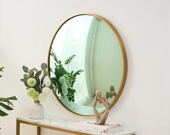 May include: A round mirror with a gold-colored frame hangs on a white wall. The mirror reflects a room with green plants. Below the mirror is a white marble-topped table with a gold frame. A small sculpture and a vase of flowers are on the table.