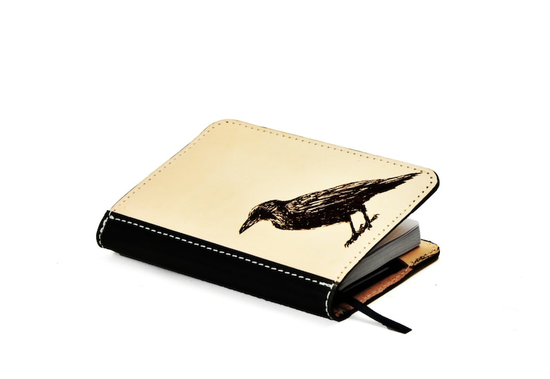 May include: A black leather journal with a cream colored cover featuring a black and white illustration of a crow. The journal is closed and the cover is slightly open.