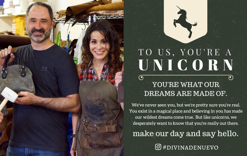May include: A couple working on leather goods in a workshop. The woman is wearing a brown leather apron and a plaid shirt. The man is holding a small, brown leather bag and a mallet. The text on the wall reads "To us, you're a unicorn. You're what our dreams are made of. We've never seen you, but we're pretty sure you're real. You exist in a magical place and believing in you has made our wildest dreams come true. But like unicorns, we desperately want to know that you're really out there. Make our day and say hello. #DIVINADENUEVO"