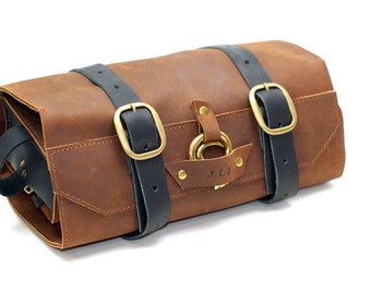 May include: A brown leather wash bag with black straps and brass buckles. The bag has a brass ring and a personalised name tag that reads "J.L.J."