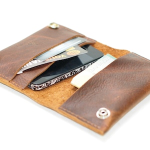 Puede incluir: Una cartera de cuero marrón con cierre de solapa y múltiples ranuras para tarjetas. La cartera está abierta y contiene un teléfono inteligente, tarjetas de crédito y efectivo.