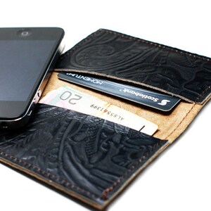 May include: A black leather wallet with an embossed design. The wallet is open and shows a credit card, cash, and a smartphone.