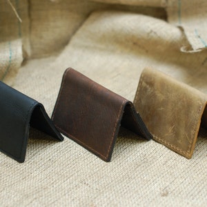 May include: Three leather card holders in black, brown, and tan, folded and displayed on a textured beige surface.
