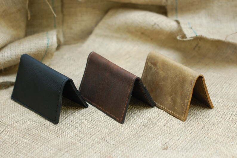 May include: Three leather card holders in black, brown, and tan, folded and displayed on a textured beige surface.