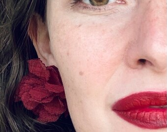 Puede incluir: Primer plano de la cara de una mujer con lápiz labial rojo y un pendiente en forma de flor de color burdeos.