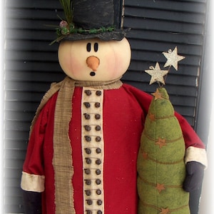 May include: A handcrafted snowman figure with a red coat, tan scarf, and black top hat. The snowman holds a green felt Christmas tree adorned with star accents. The hat is decorated with a miniature scene.