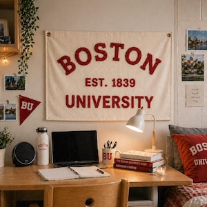 Op de afbeelding: Een crèmekleurige stoffen wandbanner met de rode tekst BOSTON EST. 1839 UNIVERSITY. De banner hangt boven een houten bureau met een laptop, een witte lamp, een notitieboek, een mok met BU-tekst en een drinkfles met Boston University branding.